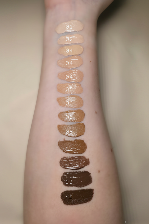 An arm with various shades of full coverage concealer applied in a line, each shade sample is labeled with a number from 01 to 13, representing different skin tones and undertones.