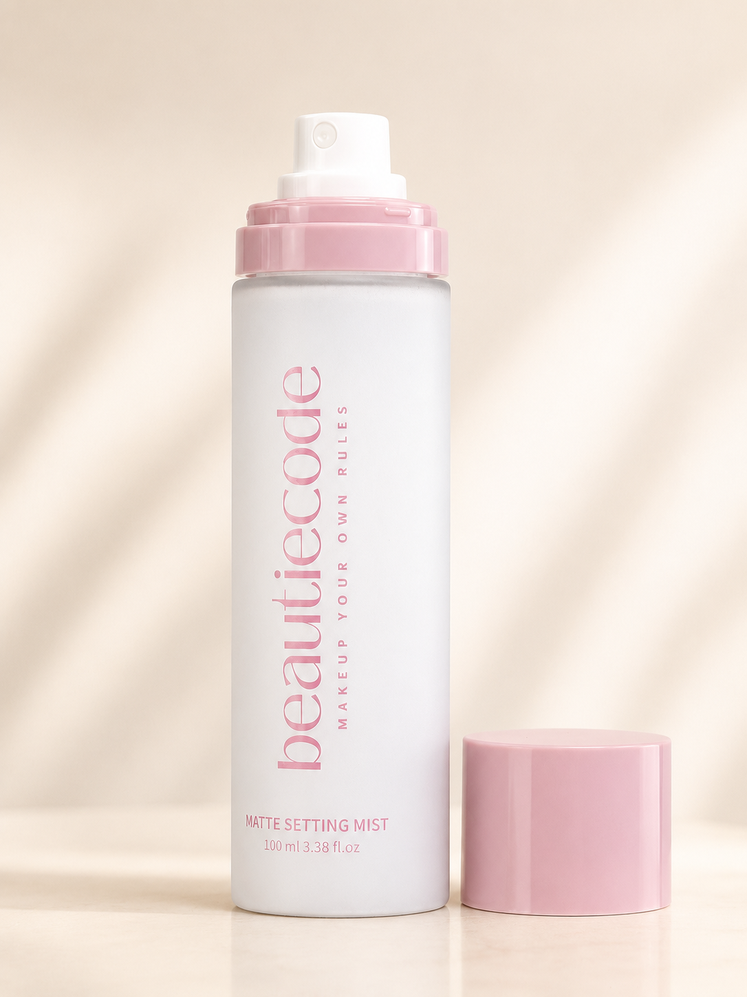 A white bottle of beautiecode Matte Setting Mist with a pink pump and a separate pink cap, set against a warm beige background with soft diagonal shadows.