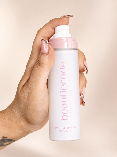 A hand with manicured nails holds a white bottle of beautiecode Matte Setting Mist against a soft beige background.