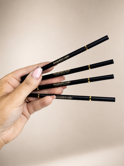 A hand with light pink manicured nails holds four black cosmetic pencils with gold accents and the brand name 'beautiecode' printed in gold on the barrels, against a solid beige background.