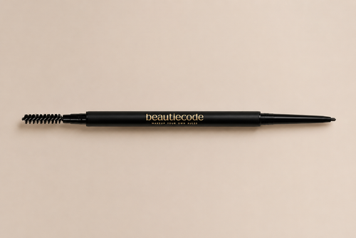 A black dual-ended eyebrow pencil with a spoolie brush on one end and a fine tip on the other, featuring the brand name 'beautiecode' in gold lettering, set against a solid beige background.