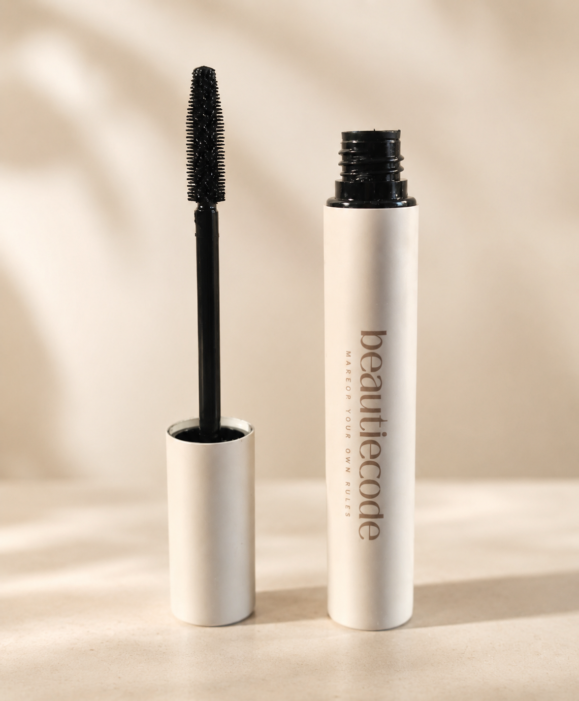 A cosmetic mascara tube shown open with the wand removed, standing beside the cap on a warm beige surface. The tube is white with black text reading 'beautiecode', and the wand is black.