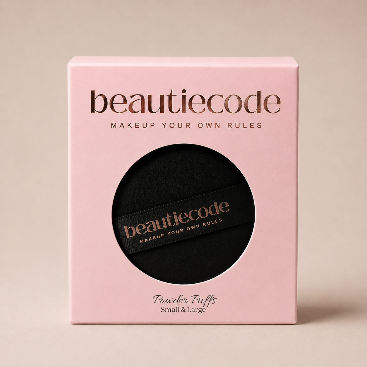 A square pink beauty box with a circular cutout showing a black makeup puff inside. The box and the puff both display the brand name 'beautiecode' in metallic rose-gold lettering, with 'Powder Puffs Small & Large' printed below on the box.