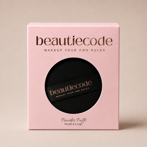 A square pink beauty box with a circular cutout showing a black makeup puff inside. The box and the puff both display the brand name 'beautiecode' in metallic rose-gold lettering, with 'Powder Puffs Small & Large' printed below on the box.