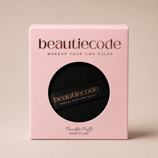 A square pink beauty box with a circular cutout showing a black makeup puff inside. The box and the puff both display the brand name 'beautiecode' in metallic rose-gold lettering, with 'Powder Puffs Small & Large' printed below on the box.