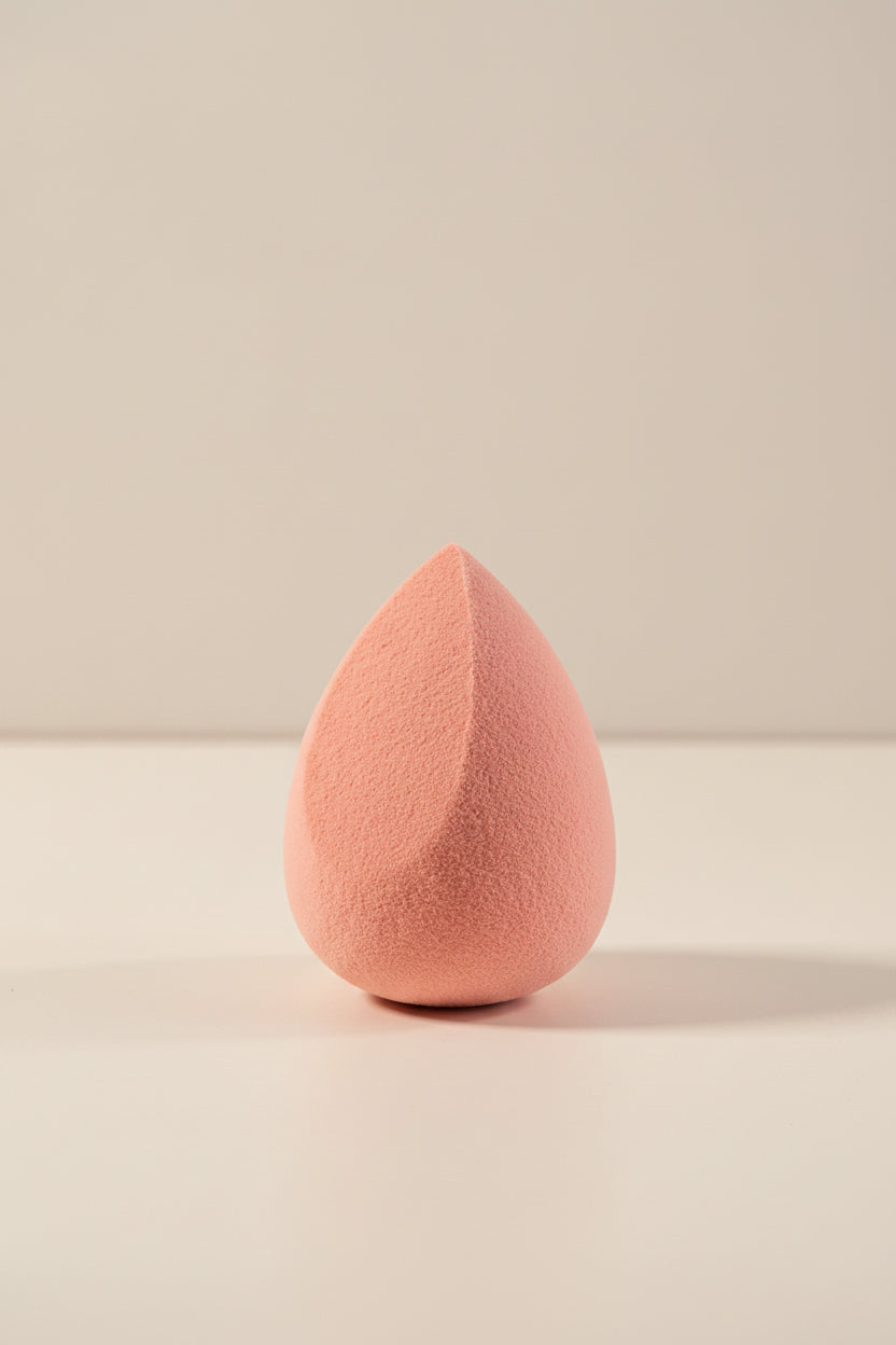 A teardrop-shaped makeup sponge in a soft dusty pink color, standing upright on a clean white surface against a light gray background.