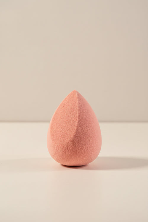 A teardrop-shaped makeup sponge in a soft dusty pink color, standing upright on a clean white surface against a light gray background.